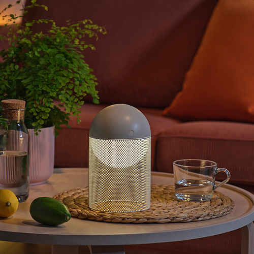 Una lámpara moderna sobre una mesa, con una planta, una taza y una fruta cerca.