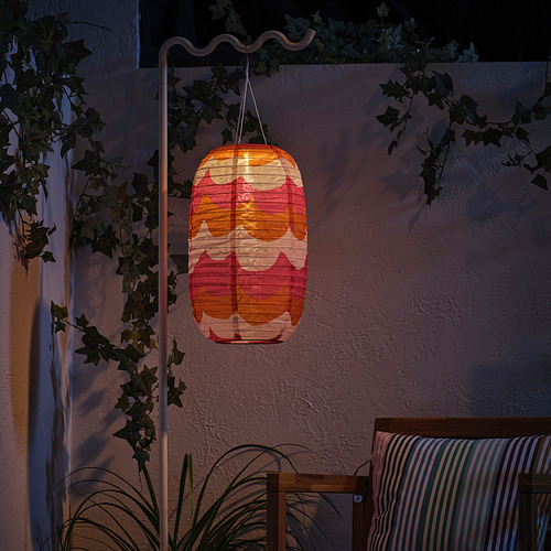 La linterna encendida cuelga de un soporte blanco, proyectando colores cálidos contra una pared texturizada, rodeada de plantas verdes.