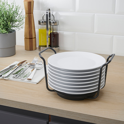 Platos blancos apilados en soporte negro sobre encimera de madera, con cubiertos y utensilios de cocina cerca.