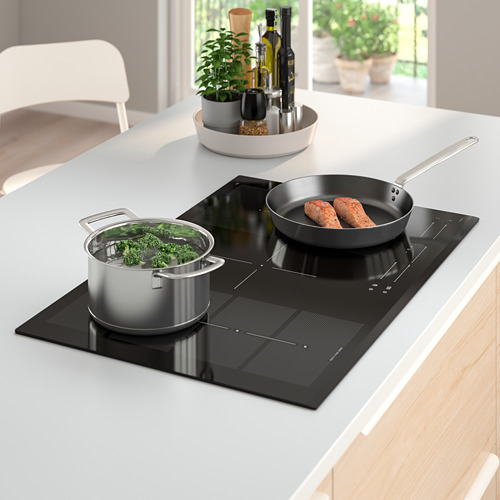 Cocina moderna con placa de inducción BLIXTSNABB, cinco zonas de cocción de verduras y embutidos. Bandeja con botellas y planta al lado.