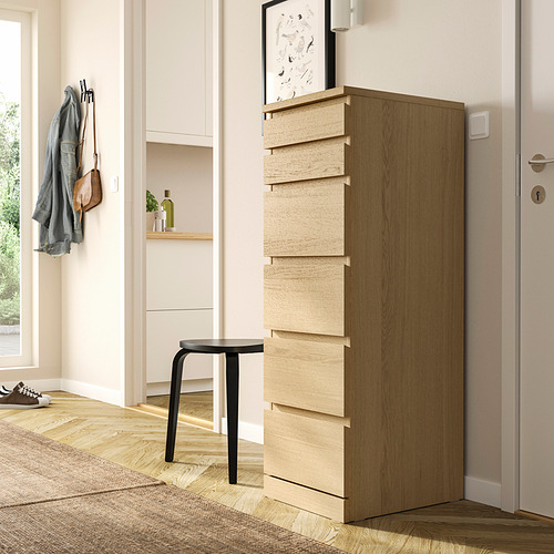 Una cómoda de madera MALM, más alta que el taburete negro cercano. Tiene seis cajones con un elegante acabado en roble marrón claro y un espejo en la parte superior.