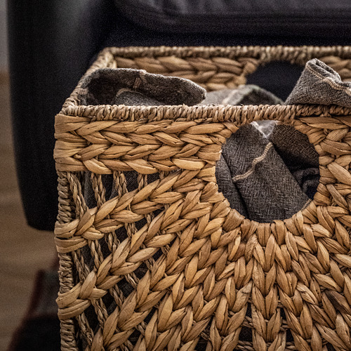 Cesta de mimbre, marrón, rectangular, rellena de artículos de color gris oscuro.
