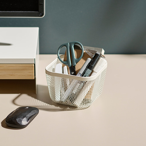 Escritorio con cesta de malla metálica blanca que sostiene tijeras, bolígrafos y regla, junto a un mouse de computadora negro y un monitor sobre un fondo verde claro.
