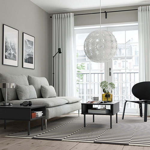 Sala de estar moderna con sofá gris, mesa de centro negra, alfombra a rayas, lámpara de pie, gran lámpara colgante y vista al balcón.