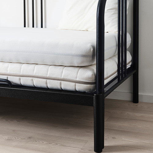 Estructura de cama de metal negro con colchón blanco y tres almohadas sobre suelo de madera.