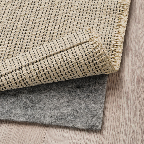 Una alfombra tejida de color beige con detalles oscuros, colocada sobre una almohadilla antideslizante gris sobre un piso de madera.