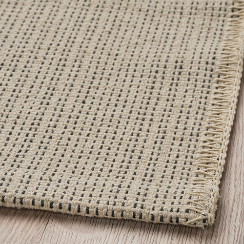 Alfombra de algodón de tejido plano, de color beige con pequeños puntos oscuros, cuidadosamente bordeada, colocada sobre suelo de madera.