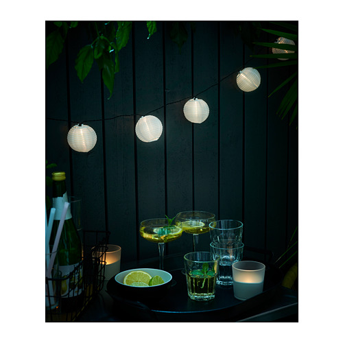 Las guirnaldas de luces iluminan un patio por la noche. Vasos, rodajas de lima y velas descansan sobre una mesa contra una valla de madera.
