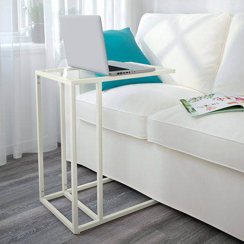 Una sala de estar moderna, un soporte para computadora portátil VITTSJÖ blanca con una tapa de vidrio, sosteniendo una computadora portátil abierta junto a un sofá blanco con cojines azules.