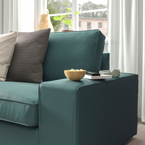 Sofá verde azulado con almohadas texturizadas, reposabrazos grande con bocadillos, control remoto y auriculares, cerca de una ventana con cortinas.
