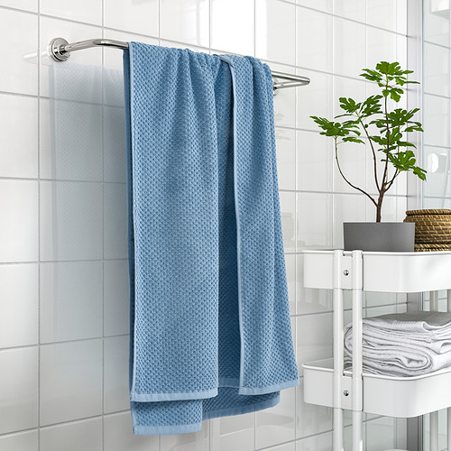 La toalla azul GULVIAL cuelga de la barra del baño, junto a estantes blancos que sostienen toallas dobladas y una planta verde.