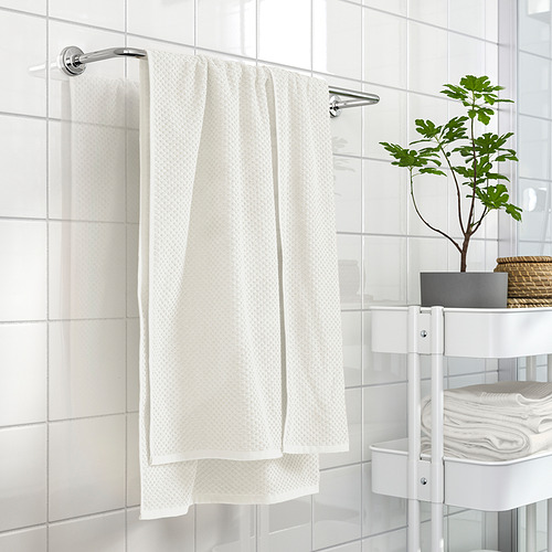 Toalla GULVIAL beige sobre varilla cromada junto a pared alicatada en blanco, con estantería y planta que muestran su tamaño y utilidad.