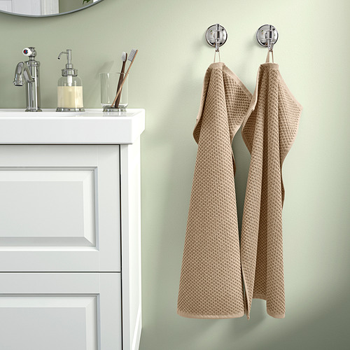 Dos toallas GULVIAL beige cuelgan junto a un lavabo blanco. Las toallas son suaves, hechas de 100% algodón, y de grosor medio, pareciendo más grandes que el lavabo.