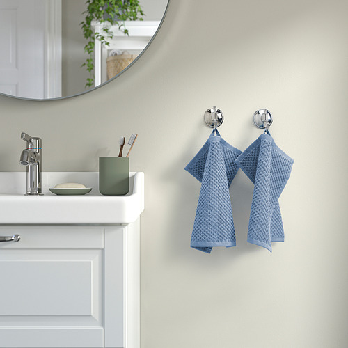 Baño: lavabo blanco, grifo cromado, taza verde. Las toallas azules cuelgan de ganchos. Espejo arriba, paredes beige.
