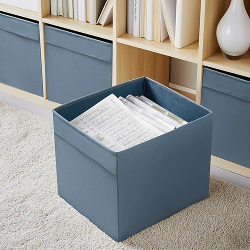 Caja de almacenamiento DRÖNA azul con papeles sobre alfombra beige, más pequeña que la estantería cercana y el jarrón decorativo.
