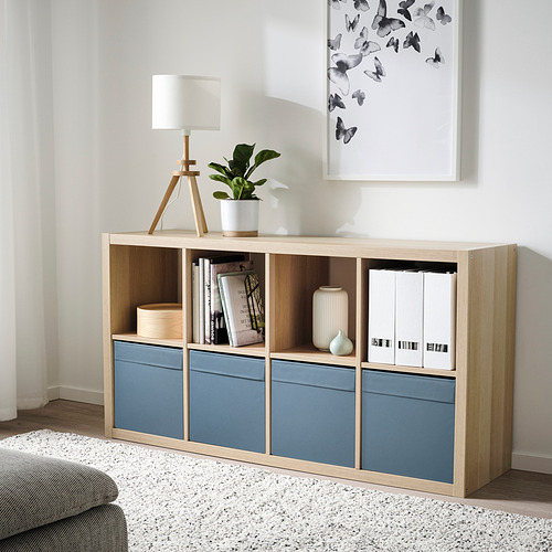Espacio habitable minimalista con unidad de almacenamiento de madera clara y azul. Contiene libros, una lámpara y plantas. La caja es estrecha, encaja debajo de la obra de arte.