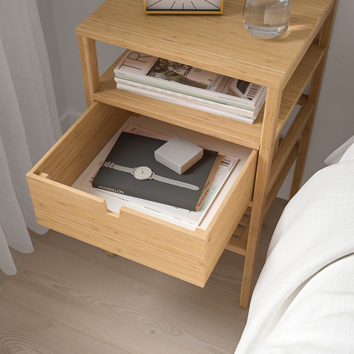 Mesita de noche de madera con cajón abierto que muestra libros, vidrio y reloj en la parte superior. El cajón tiene manijas en ambos lados.