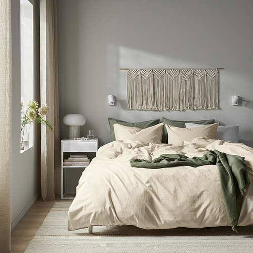 Dormitorio neutro con ropa de cama de algodón, tapices de macramé, detalles en verde, lámpara blanca, cortinas transparentes.