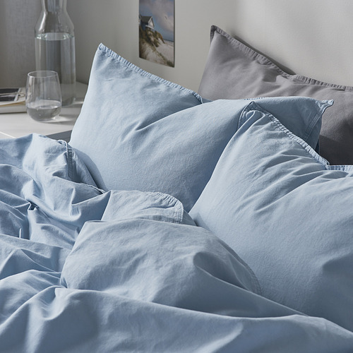 Cama con funda nórdica y almohadas azul claro, cuidadosamente dispuestas, mostrando un tejido de algodón suave y transpirable.