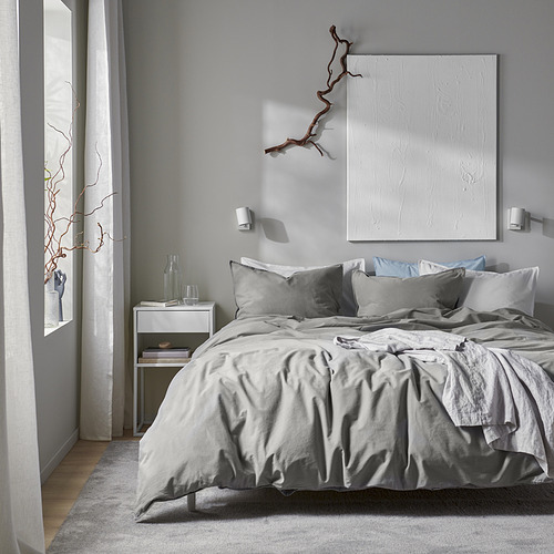 Dormitorio moderno y minimalista con funda nórdica y almohadas grises de ÄNGSLILJA, obras de arte blancas, mesita de noche plateada y decoración de ramas.