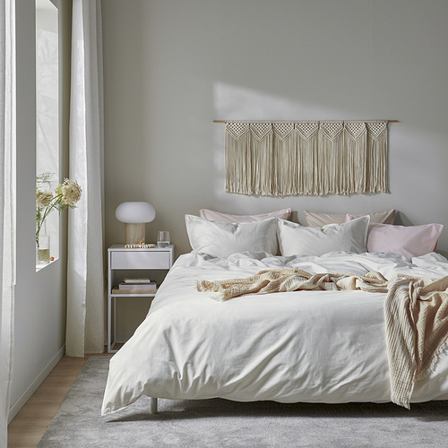 Acogedor dormitorio con edredón ÄNGSLILJA, almohadas, decoración de pared tejida, lámpara de noche, libros y flores secas. Colores suaves, centrados en la comodidad.