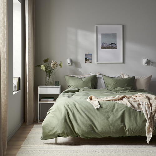 Dormitorio minimalista con juego de cama ÄNGSLILJA verde salvia. Edredón y almohadas de algodón suave, arte enmarcado, mesa auxiliar con flores, gran ventanal.