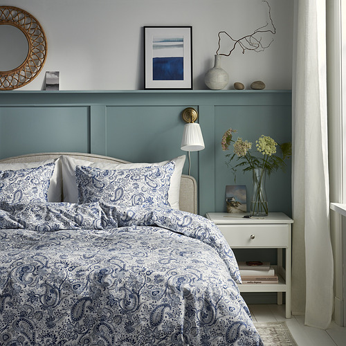 Dormitorio con cabecero azul verdoso, ropa de cama blanca, mesita de noche con libros y lámpara.