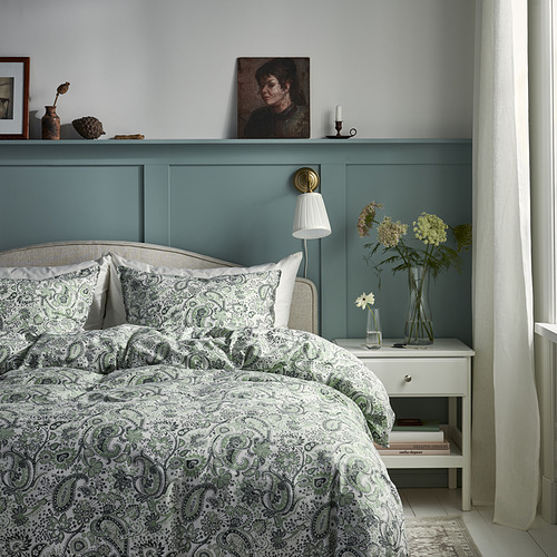 Dormitorio sereno: cama grande con edredón de cachemira verde, almohadas blancas, mesita de noche con flores, vela, retrato enmarcado y apliques de pared.