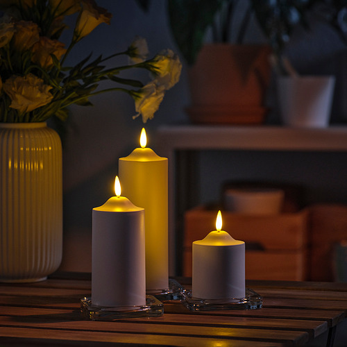 Tres velas SMÅLOM encendidas sobre la mesa, jarrón con flores amarillas, resplandor cálido.