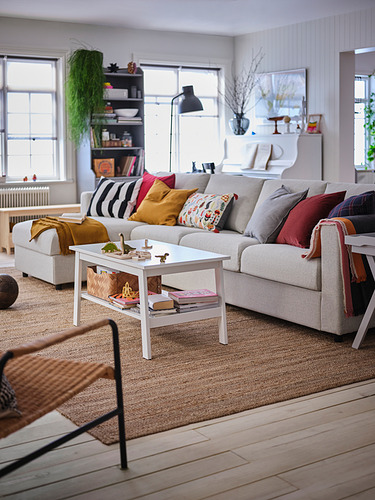 Sala de estar luminosa con sofá VIMLE gris, cojines de colores, mesa de centro blanca, grandes ventanales y plantas.