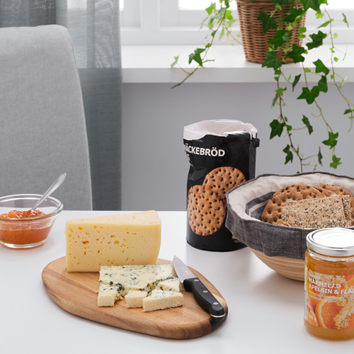 Tabla de cortar de madera con queso y cuchillo; tarro de miel, galletas saladas, mermelada y plantas.