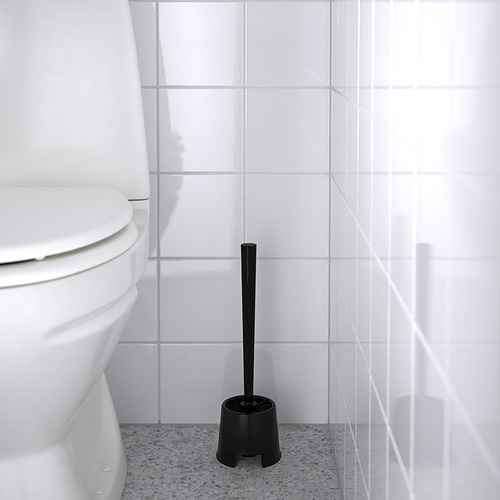 Escobilla de baño negra en soporte al lado del inodoro blanco. Herramienta de limpieza almacenada en posición vertical sobre un suelo gris moteado.