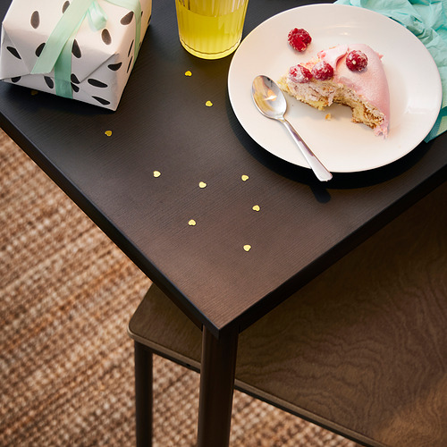 Una rebanada de pastel con frutos rojos en un plato, junto a un regalo envuelto y un vaso amarillo en una mesita negra y de madera.
