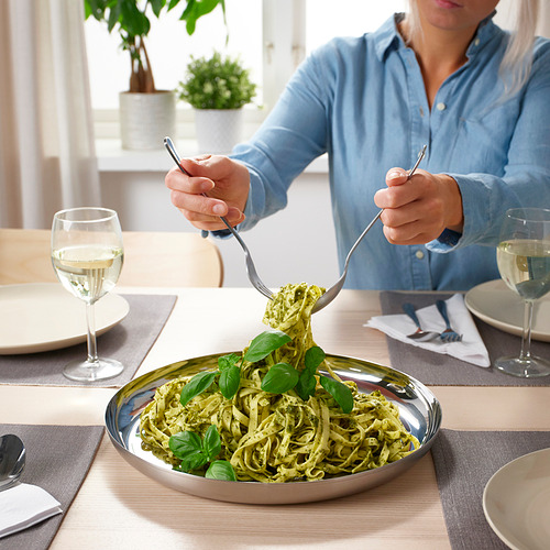 La persona hace girar la pasta con un tenedor y una cuchara alrededor de un plato redondo de acero inoxidable para servir, rodeado de copas de vino blanco y platos.