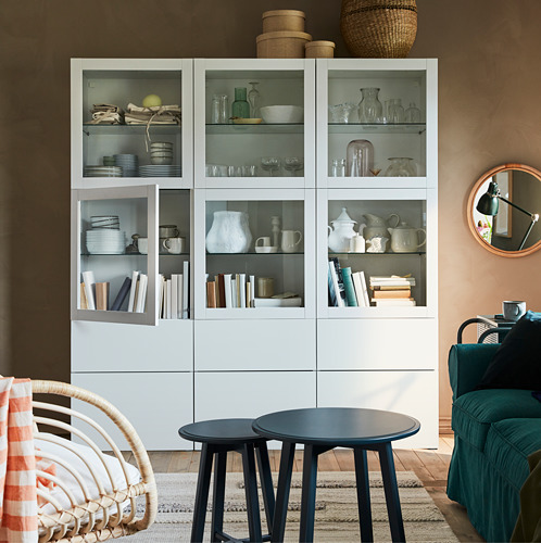 Mueble blanco BESTÅ con puertas de cristal que muestran platos y libros, mesas negras, silla de mimbre, sofá verde.