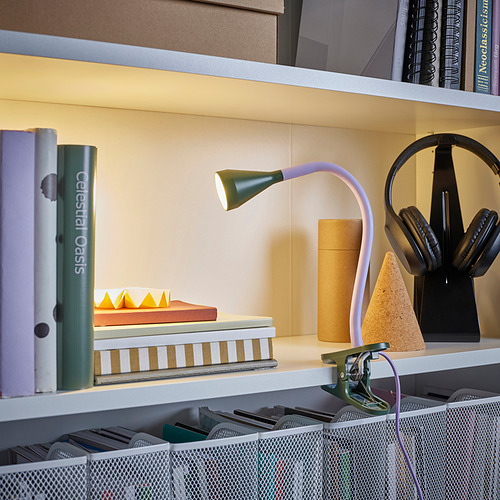 Estantería con lámpara NÄVLINGE sujeta en el borde, iluminación de libros. También se ven auriculares y varias pilas de libros.