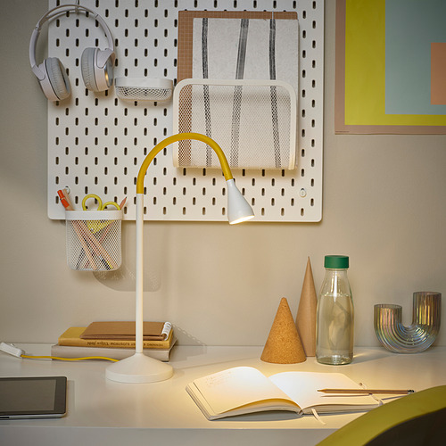 Pequeña lámpara de escritorio amarilla moderna, NÄVLINGE, sobre un escritorio luminoso con libros y una tableta. Tiene un cuello y una base flexibles, lo que proporciona una iluminación focalizada.