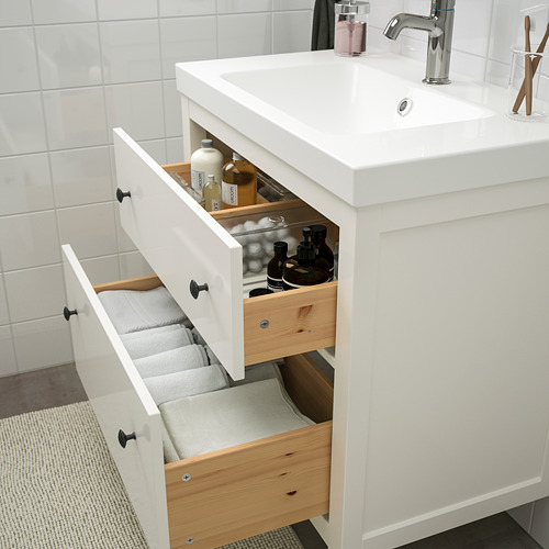 Mueble de baño blanco con cajones abiertos que muestran toallas, botellas y artículos de tocador, debajo de un lavabo con grifo cromado.