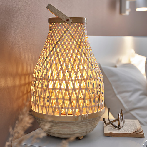Una lámpara MISTERHULT con marco de bambú en color beige natural, con una luz cálida y brillante, colocada sobre una mesita de noche.