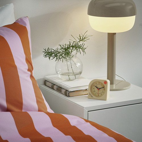 Cama a rayas con mesita de noche. La mesa tiene libros, un jarrón redondo con vegetación, un reloj con alarma luminosa y una lámpara encendida.