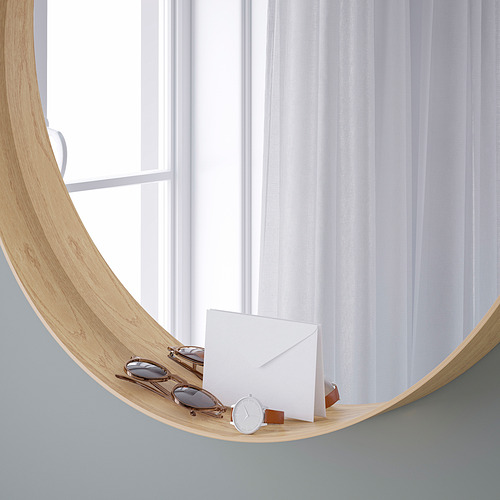 Un espejo con marco de madera y un estante inferior contiene gafas de sol, un reloj y un sobre. El espejo está colocado junto a una ventana con cortinas blancas transparentes.