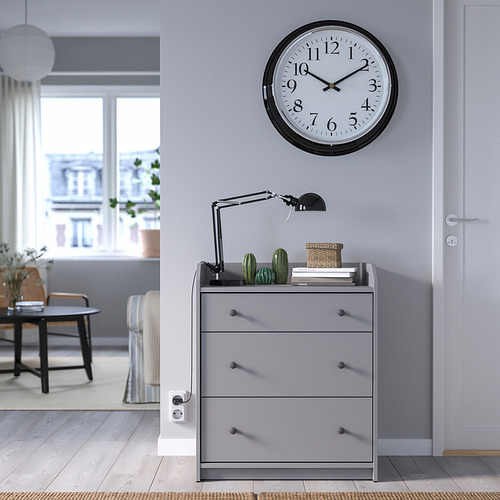 Moderno cofre gris con reloj, lámpara, cesta y plantas.