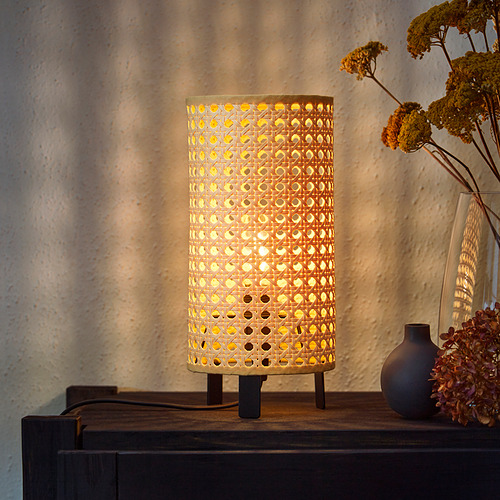 Una lámpara SAXHYTTAN con pantalla de papel perforado, que proyecta una luz cálida. Se sienta sobre una superficie de madera con flores secas en un jarrón a su lado.