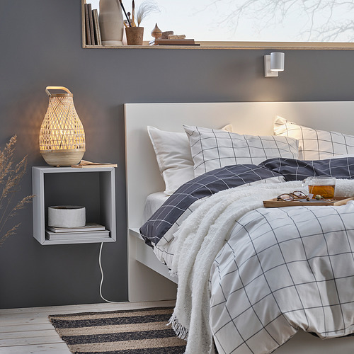 Esta imagen muestra un dormitorio sereno con una lámpara de bambú blanca MISTERHULT sobre una mesita de noche gris. La cama tiene ropa de cama blanca y gris con estampado de rejillas.