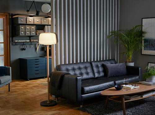 Sala de estar moderna con sofá de cuero LANDSKRONA negro, mesa de centro de madera, lámpara alta y separador de pared a rayas.