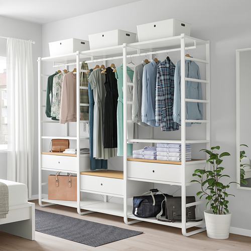 El armario blanco ELVARLI con estantes y cajones almacena varias prendas y bolsos, colocados junto a una alfombra gris y una cama blanca, con una planta a su lado.
