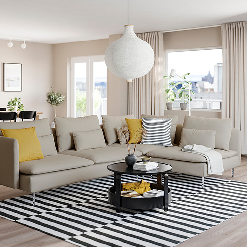Sala de estar moderna con sofá SÖDERHAMN en beige, almohadas amarillas y negras, alfombra a rayas, lámpara colgante, grandes ventanales, plantas y mesa de café.