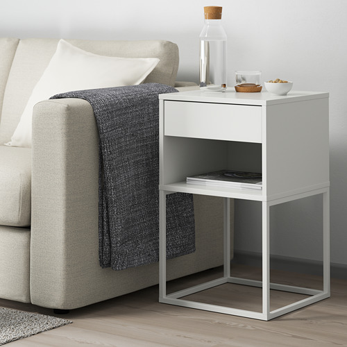 Mesa auxiliar blanca junto al sofá con cajón, estante, botella, cuencos, revistas, manta gris.