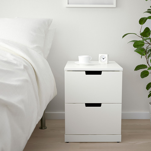 Moderna mesita de noche NORDLI blanca con dos cajones, entre la cama y la planta.