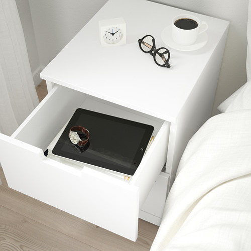 Mesita de noche con cajón abierto que muestra tableta y reloj. Reloj, vasos y café encima.